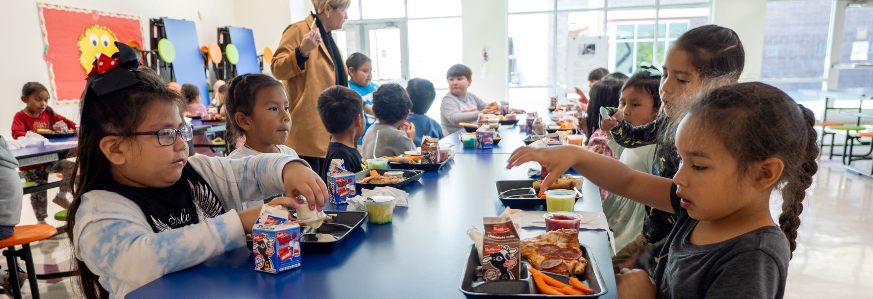 school cafeteria full of students sitting at long table with trays of food