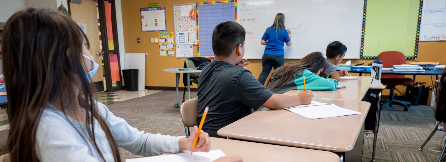 A group of students sitting at desks in a classroom, watching attentively at what the teacher is writing on the board.
