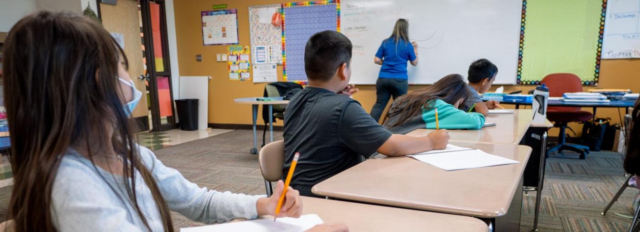 A group of students sitting at desks in a classroom, watching attentively at what the teacher is writing on the board.